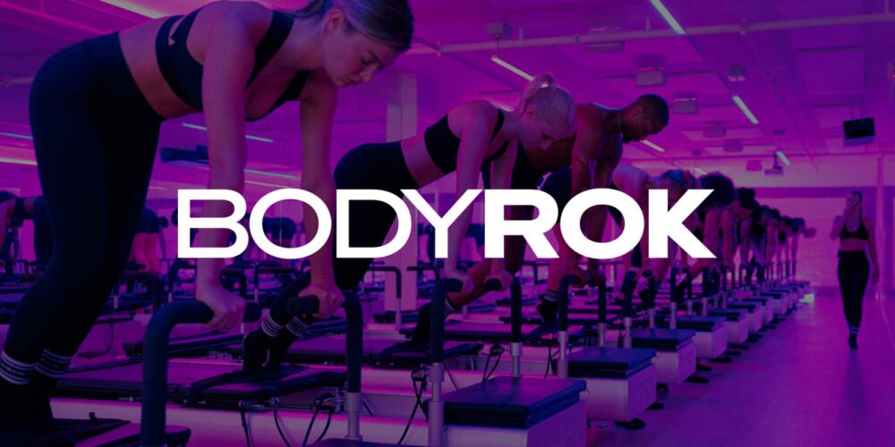 Group fitness class on reformer machines in a neon-lit studio, participants performing strength exercises while an instructor walks down the center aisle.