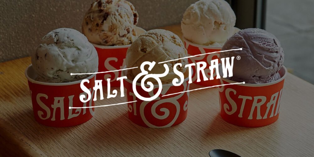 Five large scoops of Salt & Straw ice cream in red cups on a wood table next to two black spoons.